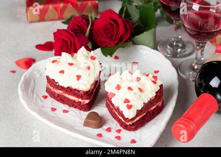 Kuchen Roter Samt in Form von Herzen auf weißem Teller, Rosen und Flaschenwein zum Valentinstag Stockfoto