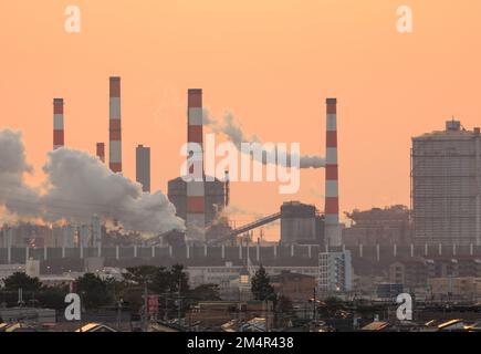 Rauch steigt aus Industriebetrieben mit orangefarbenem Sonnenschein am Himmel auf Stockfoto