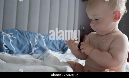 Nahaufnahme ein fröhliches, verspieltes Kind im Alter von 10 Monat. Ein neugeborener Junge beißt die Fernbedienung des Fernsehers auf ein weiches Bett. Baby-Kind-Wachzeit Kindheit, Mutterschaft, Familie Stockfoto