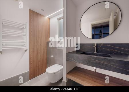 Modern gestaltete Toilette mit rundem schwarzem Spiegel, Waschbecken aus schwarzem Marmor mit Adern und weißer Hängetoilette Stockfoto