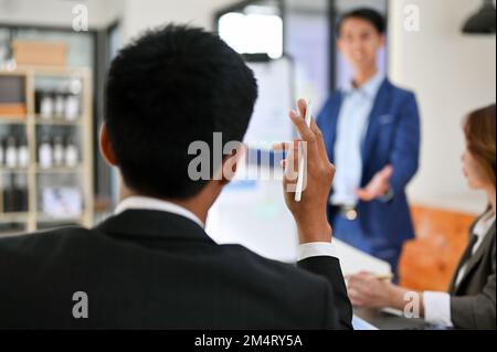 Ein kluger asiatischer Geschäftsmann, der während des Meetings mit einem Finanzanalytiker die Hand hebt und ein paar Fragen stellt. Rückansicht Stockfoto