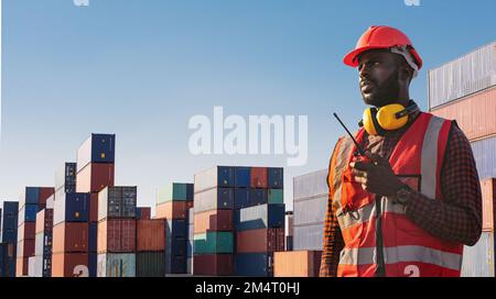 Amerikanischer afrikanischer Ingenieur oder Fabrikarbeiter, der ein Walkie-Talkie benutzt und Waren im Containerfrachthafen überprüft. Stockfoto