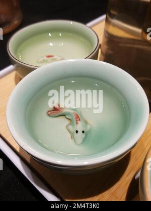 Zwei kleine, mintgrüne chinesische Teetassen (Cha bei) gefüllt mit hellem Tee, mit zartem 3D-Keramik-Koi-Fisch-Dekor am Boden. Stockfoto