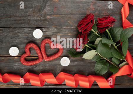 Wunderschöner roter Rosenstrauß, Kerzen und rotes Band mit Herzen auf einem Holztisch, Platz für Text. Konzept der Valentinstagsfeier Stockfoto