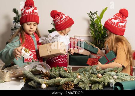 Drei Kinder in roten Weihnachtshüten packen Geschenke und machen Weihnachtsdekorationen. Das Konzept des Hobbys, der Familie, des Urlaubs und des umweltfreundlichen Lebensstils. Stockfoto