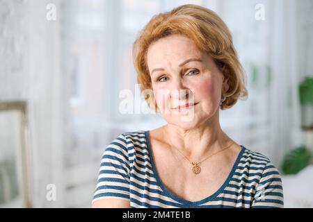 Lächelnde, graue Frau mittleren Alters, die in die Kamera schaut, glückliche alte Frau in gestreiftem T-Shirt, die daheim posiert, positive einsame ältere Frau im Schlafzimmer Stockfoto