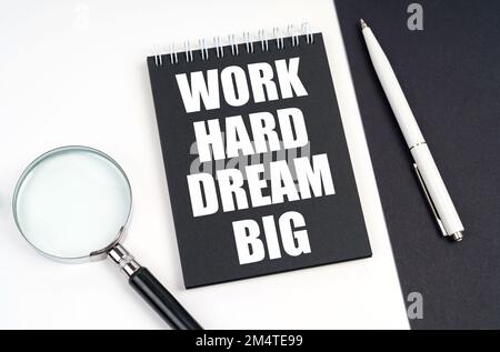 Business- und Economy-Konzept. Auf einer schwarz-weißen Oberfläche liegen eine Lupe, ein Stift und ein Notizblock mit der Aufschrift „Work Hard Dream Big“ Stockfoto