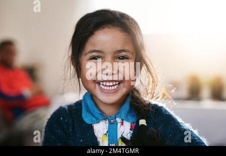 Nichts kann ihr Lächeln nehmen. Porträt eines fröhlichen kleinen Mädchens, das tagsüber zu Hause herumspielt und dabei den ganzen Weg hineinlächelt. Stockfoto