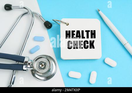 Medizinisches Konzept. Auf einer weißen und blauen Oberfläche sind Pillen, ein Stethoskop, ein Stift und ein Notizblock mit der Inschrift - HEALTH CHECK Stockfoto