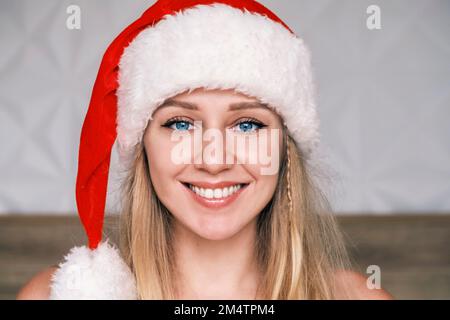 Lächelndes Weihnachtsmann-Mädchen mit rotem Hut, das in die Kamera schaut. Porträt einer schönen, blauen, blonden Frau mit Weihnachtsmannmütze. Weihnachtsporträt. Weihnachten Stockfoto