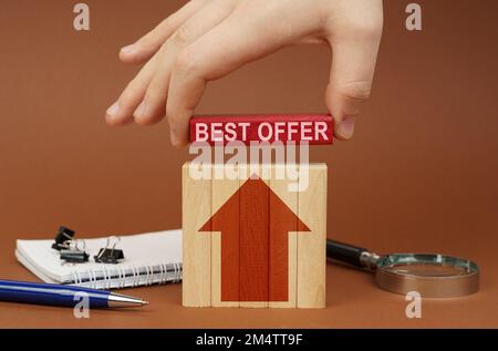 Geschäftskonzept. Auf einer braunen Oberfläche befinden sich Bürogegenstände, Holzblöcke, in der Hand befindet sich ein roter Block mit der Aufschrift "Bestes Angebot" Stockfoto