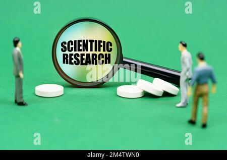 Medizinisches Konzept. Auf einer grünen Oberfläche befinden sich Pillen, Miniaturfiguren von Menschen und eine Lupe mit der Aufschrift "Wissenschaftliche Forschung" Stockfoto