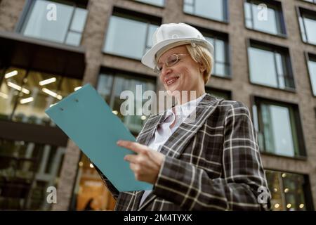 Leitende Ingenieurin im Anzug mit weißem Helm, die sich auf einem Tablet Notizen über den Gebäudehintergrund macht Stockfoto