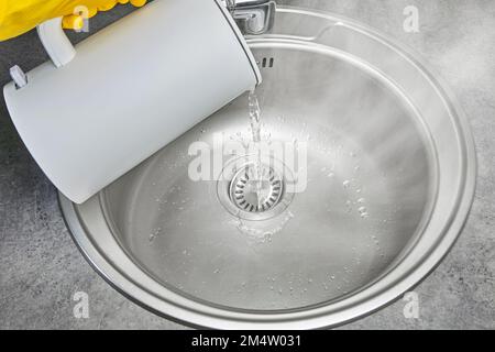 Eine Person in gelben Gummihandschuhen gießt heißes Wasser mit Dampf aus einem elektrischen Wasserkocher in ein Metallwaschbecken Stockfoto