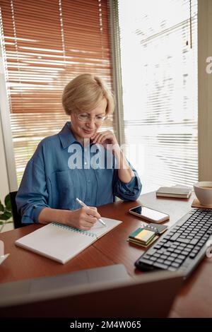 Schöne reife Frau, die sich Notizen macht, während sie im gemütlichen Home Office arbeitet Stockfoto