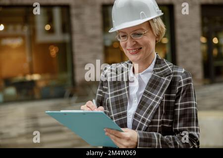 Lächelnde Ingenieurin im Anzug mit weißem Helm, die sich im Hintergrund Notizen in Tabletts macht Stockfoto