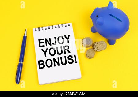 Geschäfts- und Finanzkonzept. Auf einer gelben Oberfläche, einem Sparschwein, Münzen und einem Notizblock mit der Inschrift – genießen Sie Ihren Bonus Stockfoto