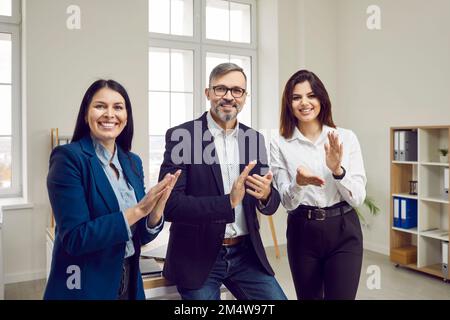 Brutaler, grauhaariger, glücklicher Mann und zwei schöne, lächelnde junge Geschäftsfrauen applaudieren, wenn sie in die Kamera schauen. Stockfoto