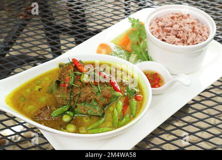 Warmes und würziges grünes thailändisches Curry mit Rindfleisch und eine Schüssel gekochten roten Thai Cargo Reis im Hintergrund Stockfoto