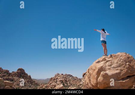 Eine Frau, die auf einem großen Felsen steht und den versteckten Valley Trail im Joshua Tree-Nationalpark im sonnigen kalifornien überblickt. Stockfoto