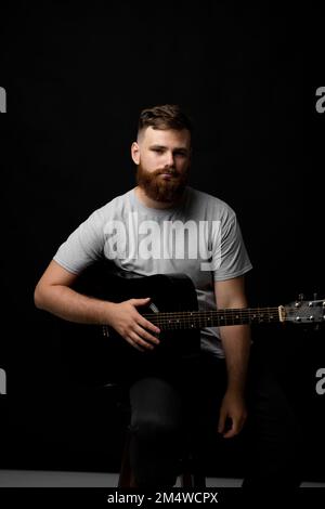 Ein bärtiger Musiker, der in einem dunklen Raum Akustikgitarre spielt. Stockfoto
