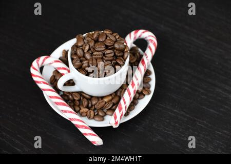 Weihnachtliche Zuckerstangen und Kaffeebohnen in einer weißen Tasse auf einem dunklen Holztisch. Dunkel gerösteter Kaffee, natürliches Getränk für Winterferien Stockfoto