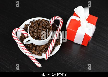 Zuckerstangen Kaffeebohnen in weißer Tasse und roter Geschenkbox auf dunklem Holztisch. Dunkel gerösteter Kaffee, natürliches Getränk für Neujahrsferien Stockfoto