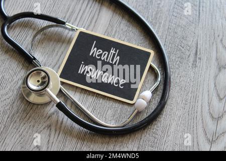 Stethoskop und Tafel mit Text KRANKENVERSICHERUNG auf Holztisch. Medizin- und Gesundheitskonzept Stockfoto