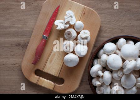 Frischer Champignon auf dem Tisch, in einer Schüssel und auf einem Holzbrett mit einem roten Messer, Draufsicht auf die Gemüsezutaten zum Kochen. Stockfoto