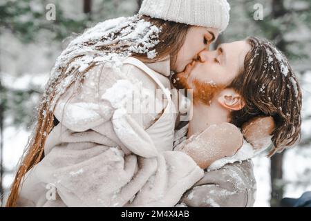 Romantische Schnee-Liebesgeschichte. Junge Frau, die im schneebedeckten Winterwald mit Bäumen küsst. Spazieren, Spaß haben, in stilvollen warmen Kleidern lachen Stockfoto
