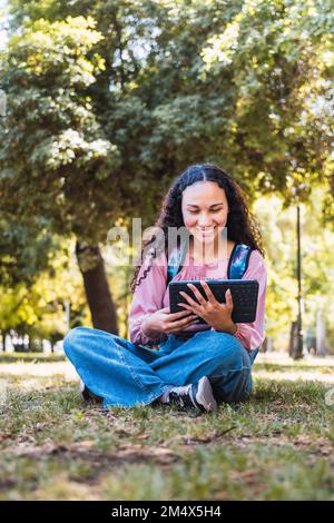 Schwarze Studentin, die lächelt und ein Tablet benutzt, das draußen in einem Park auf dem Rasen sitzt Stockfoto