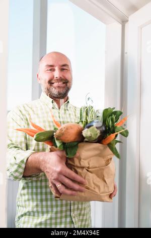 Lächelnder Mann, der frisches Bio-Gemüse in einer Papiertüte nahe der Tür hält Stockfoto