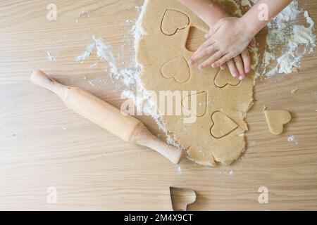 Hände von Mädchen, die Keksteig schneiden, mit Schneidevorrichtung auf dem Tisch zu Hause Stockfoto