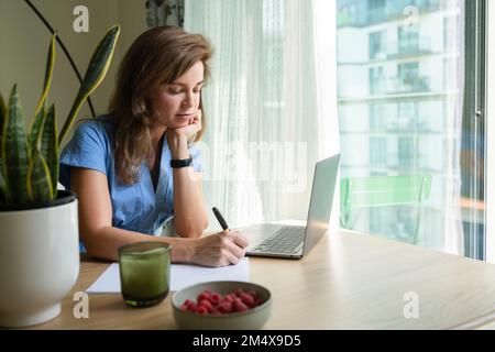 Geschäftsfrau mit Hand am Kinn, die am Schreibtisch im Heimbüro auf Papier schreibt Stockfoto