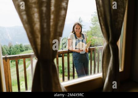 Glückliche junge Frau mit einem Buch, das durch das Fenster gesehen wurde Stockfoto