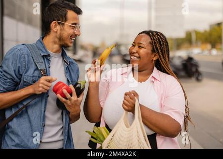 Ein fröhlicher junger Mann mit einer Freundin, die einen Sack Gemüse auf dem Fußweg hält Stockfoto