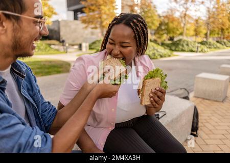 Ein glücklicher Mann, der ein Sandwich mit einer Frau teilt, die auf dem Platz am Fußweg sitzt Stockfoto