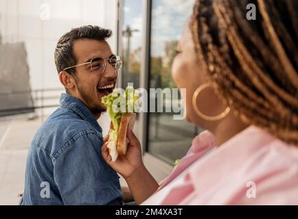 Glückliche junge Frau, die sich ein Sandwich mit einem Mann auf dem Fußweg teilt Stockfoto