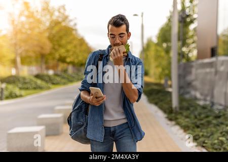 Junger Mann, der Sandwich isst und sein Smartphone auf dem Fußweg benutzt Stockfoto