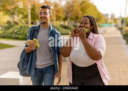 Glückliches junges Paar, das Sandwich aß und auf dem Fußweg spazierte Stockfoto