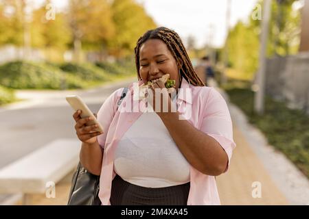 Junge Frau, die ein Smartphone benutzt und Sandwich isst, steht auf dem Fußweg Stockfoto