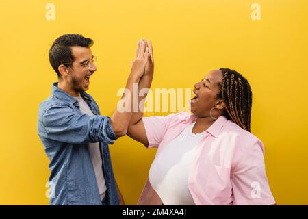 Fröhlicher Mann und Frau, die sich vor der gelben Wand gegenseitig High Five geben Stockfoto