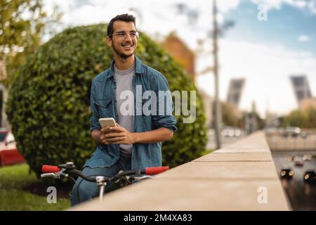 Glücklicher junger Mann mit Smartphone, der auf dem Fahrrad sitzt Stockfoto