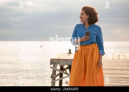 Lächelnde Frau, die mit dem Handy auf dem Steg steht, bei Sonnenuntergang Stockfoto