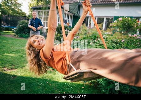 Fröhliche Frau, die auf der Schaukel schwingt, mit einem Mann, der vor dem Haus steht Stockfoto