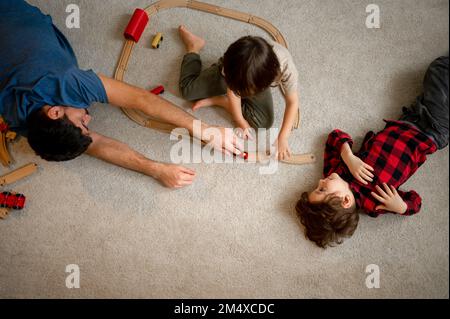 Ein Junge, der Vater und Bruder ansieht und mit einem hölzernen Spielzeugzug auf dem Boden spielt Stockfoto