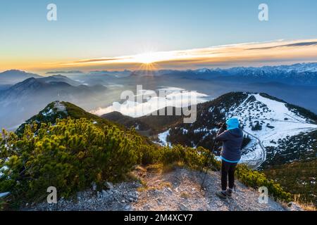 Deutschland, Bayern, Frau, die die umliegende Landschaft bei Sonnenaufgang vom Gipfel des Herzogstands fotografiert Stockfoto