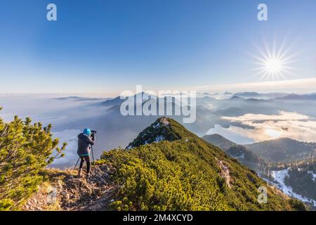 Deutschland, Bayern, Frau, die die umliegende Landschaft bei Sonnenaufgang vom Gipfel des Herzogstands fotografiert Stockfoto