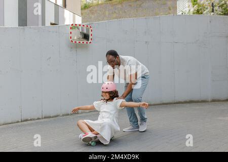 Vater schubst Tochter, die vor der Wand auf dem Skateboard sitzt Stockfoto