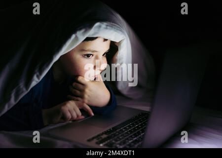 Ein rücksichtsvoller Junge, der auf dem Bett unter der Decke auf dem Laptop liegt Stockfoto